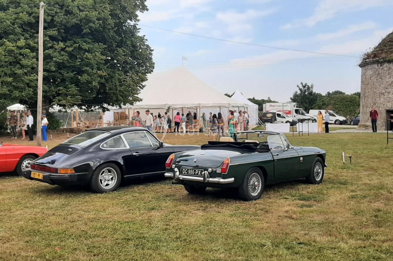 Voitures anciennes garées sur une pelouse près d'une tente blanche et d'une petite tour en pierre, par temps clair.