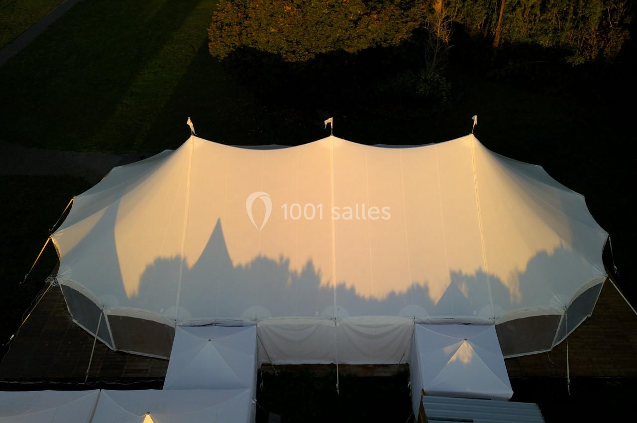 Vue aérienne d'une grande tente blanche installée sur une pelouse, entourée d'arbres en arrière-plan.