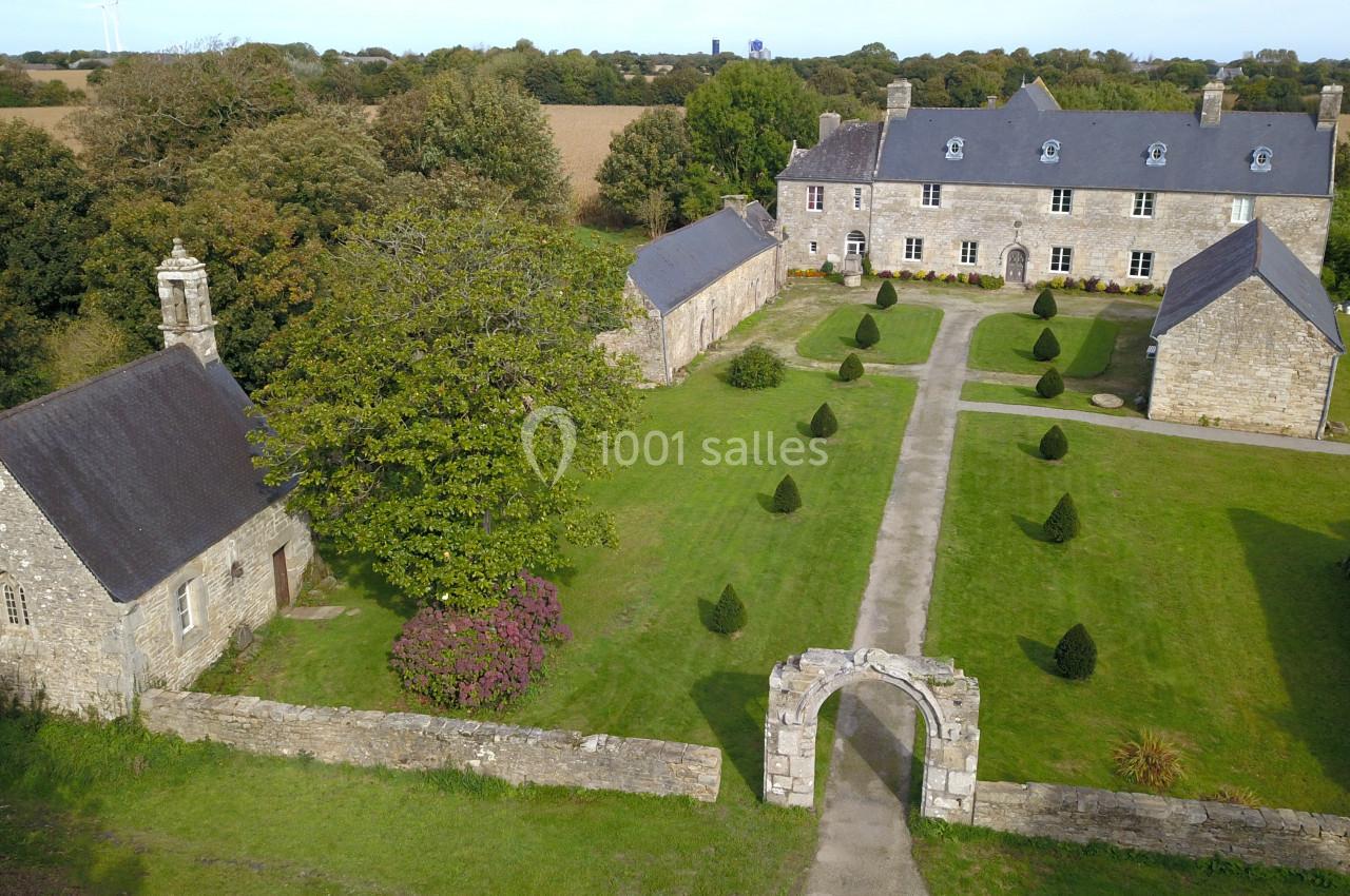 Vue aérienne d'un manoir en pierre avec une chapelle, un jardin aménagé et des champs en arrière-plan.