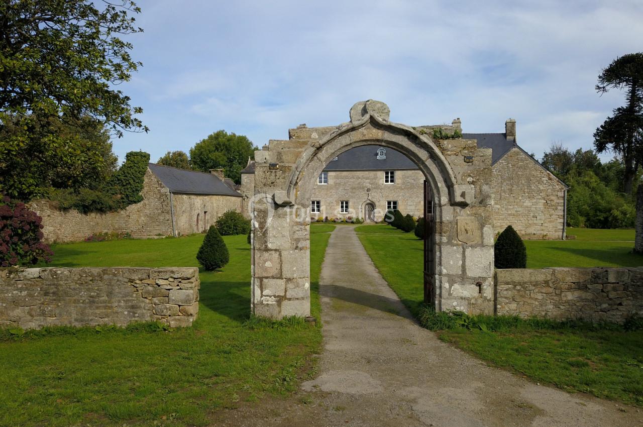 Portail en pierre ancienne menant à une maison en pierre entourée de verdure et d'arbres.
