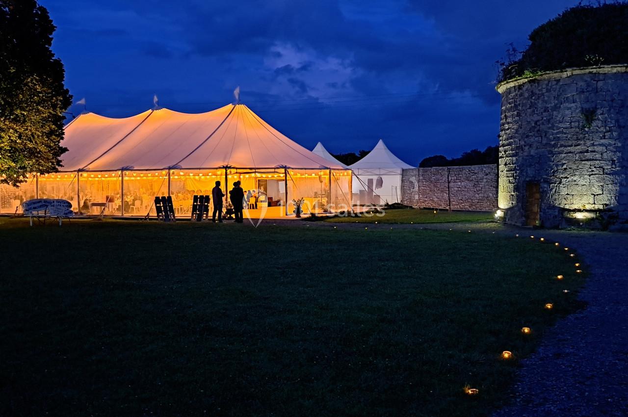 Tente éclairée en soirée dans un jardin, avec un chemin bordé de bougies menant à une ancienne tour en pierre.