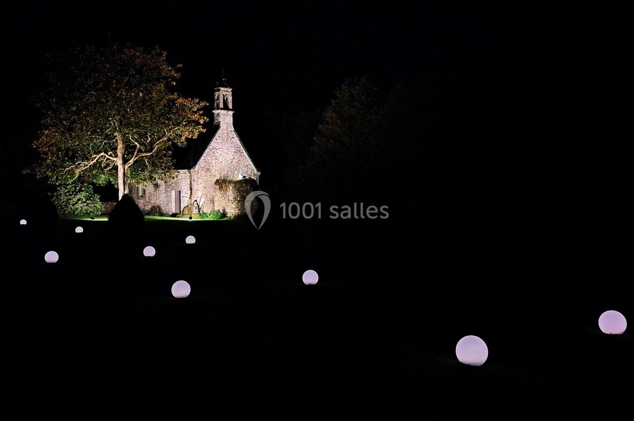 Petite chapelle en pierre éclairée la nuit, entourée d'arbres et de sphères lumineuses dans un jardin sombre.