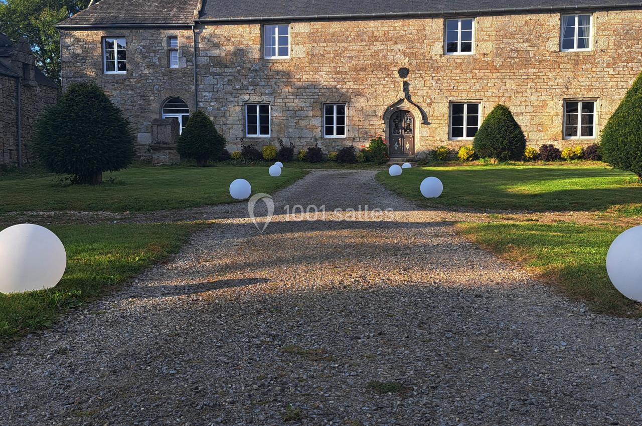 Allée en gravier bordée de pelouses et de sphères lumineuses blanches menant à un bâtiment en pierre.