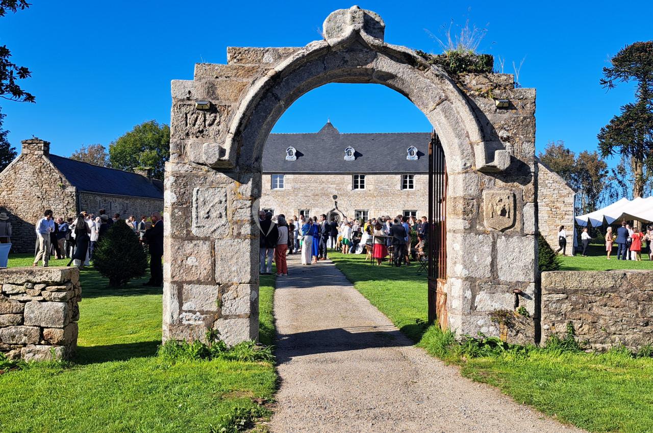 Portail en pierre donnant sur une cour avec des bâtiments anciens, des invités et des tentes blanches en arrière-plan.