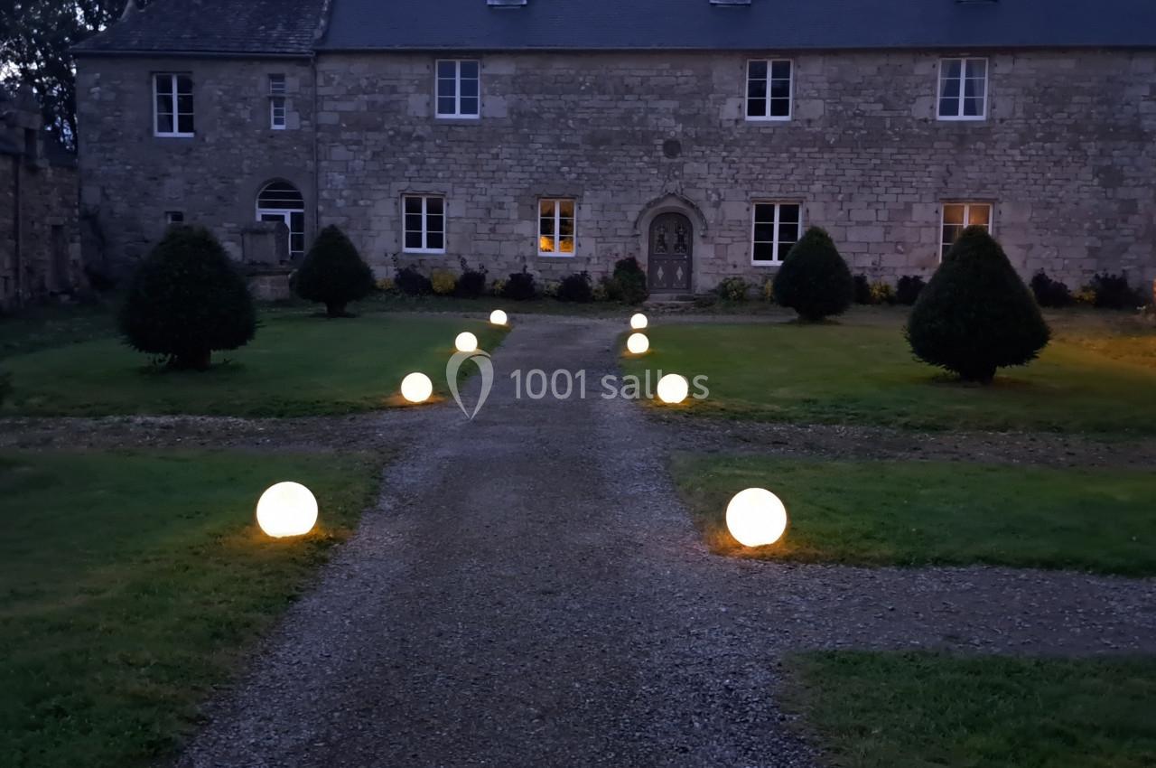 Allée gravillonnée éclairée par des sphères lumineuses, menant à une grande bâtisse en pierre au crépuscule.