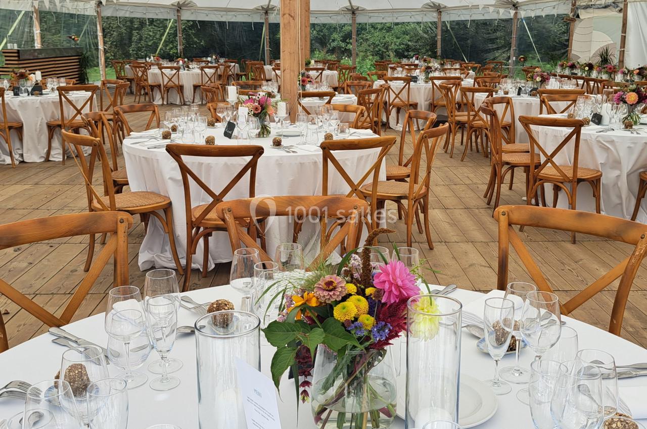 Salle de réception sous chapiteau avec tables rondes dressées, nappes blanches et bouquets de fleurs colorées.
