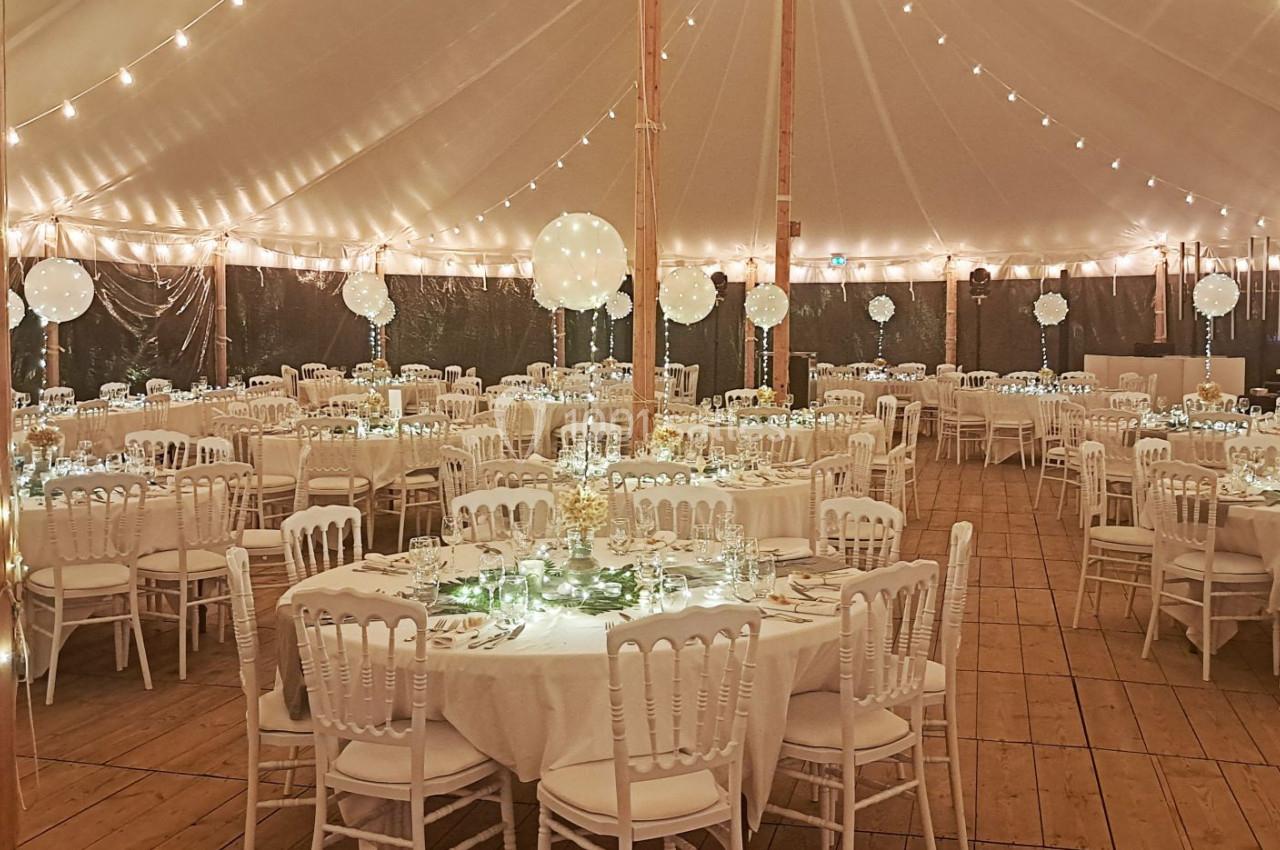 Salle de réception sous une tente éclairée, avec des tables rondes décorées et des chaises blanches alignées.