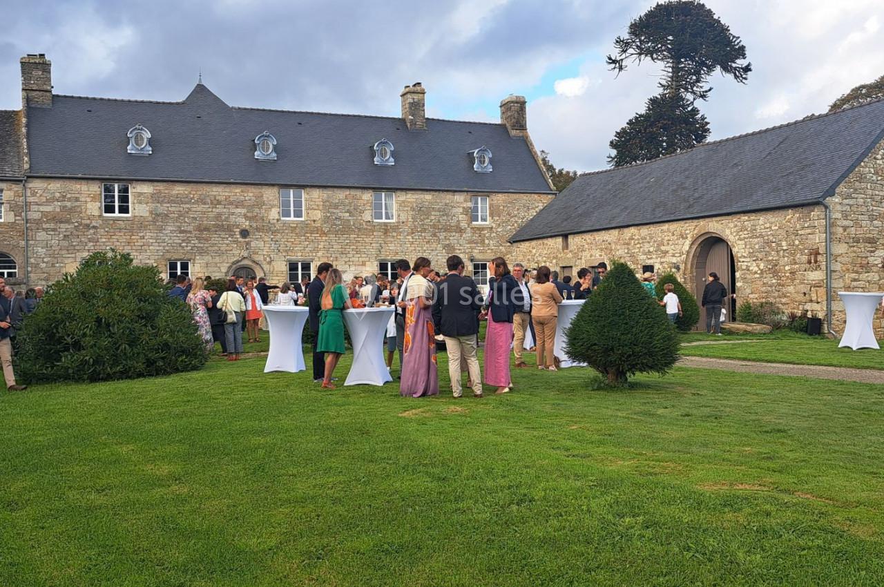 Groupe de personnes rassemblées dans un jardin devant un bâtiment en pierre, lors d'un événement en plein air.