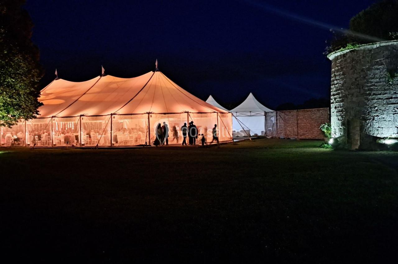 Tente éclairée en soirée avec des silhouettes à l'intérieur, entourée d'un espace vert et d'une tour en pierre.