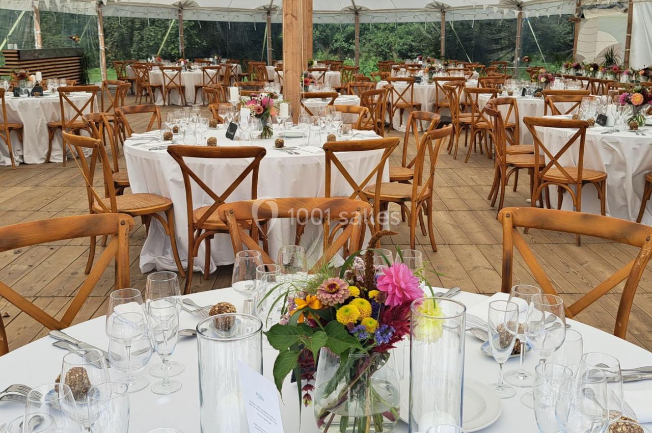 Salle de réception sous une tente avec tables rondes dressées, chaises en bois et décorations florales colorées.