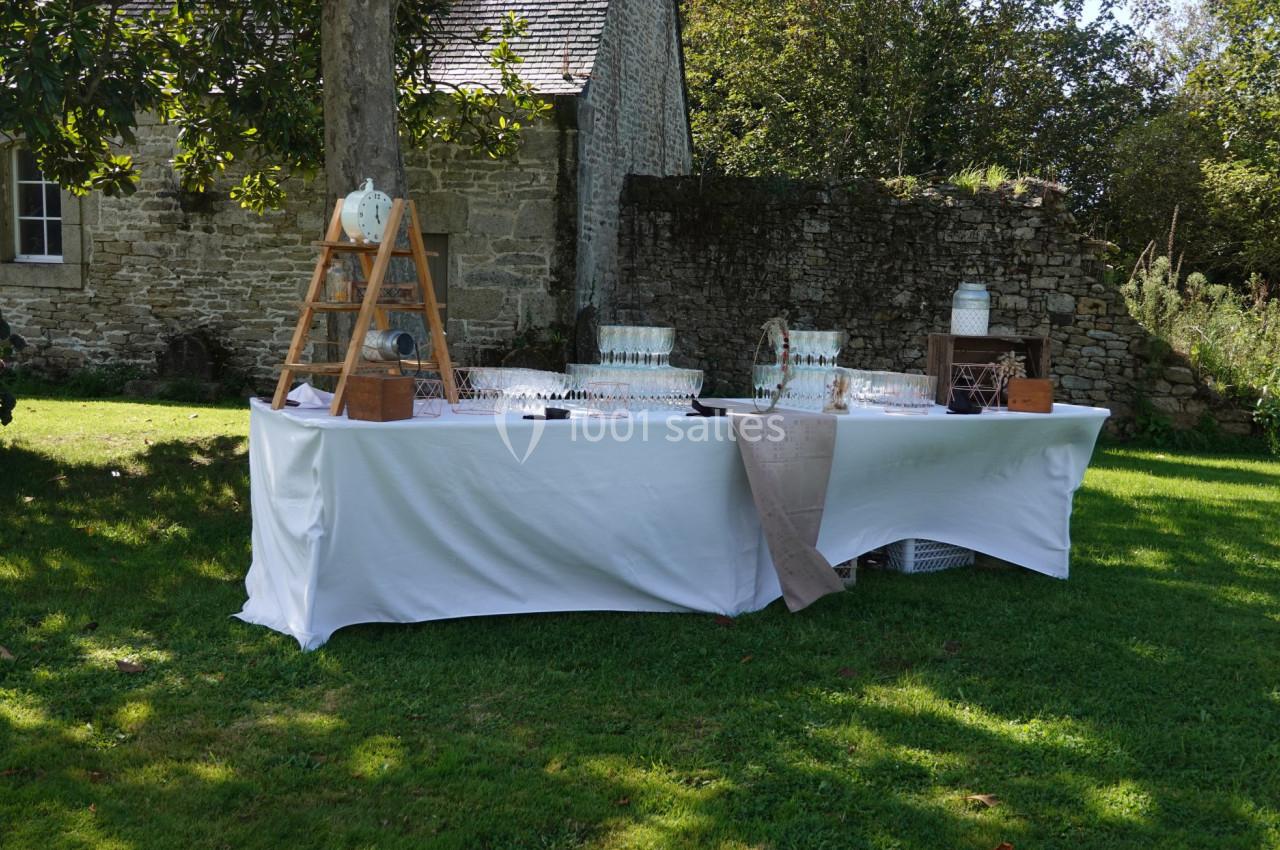 Table dressée en extérieur avec nappes blanches, verres alignés et décorations rustiques près d'un mur en pierre.