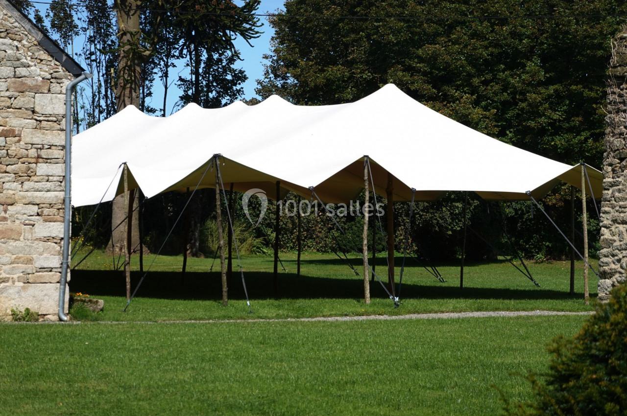 Tente blanche montée sur un espace herbeux, entourée d'arbres et de murs en pierre.