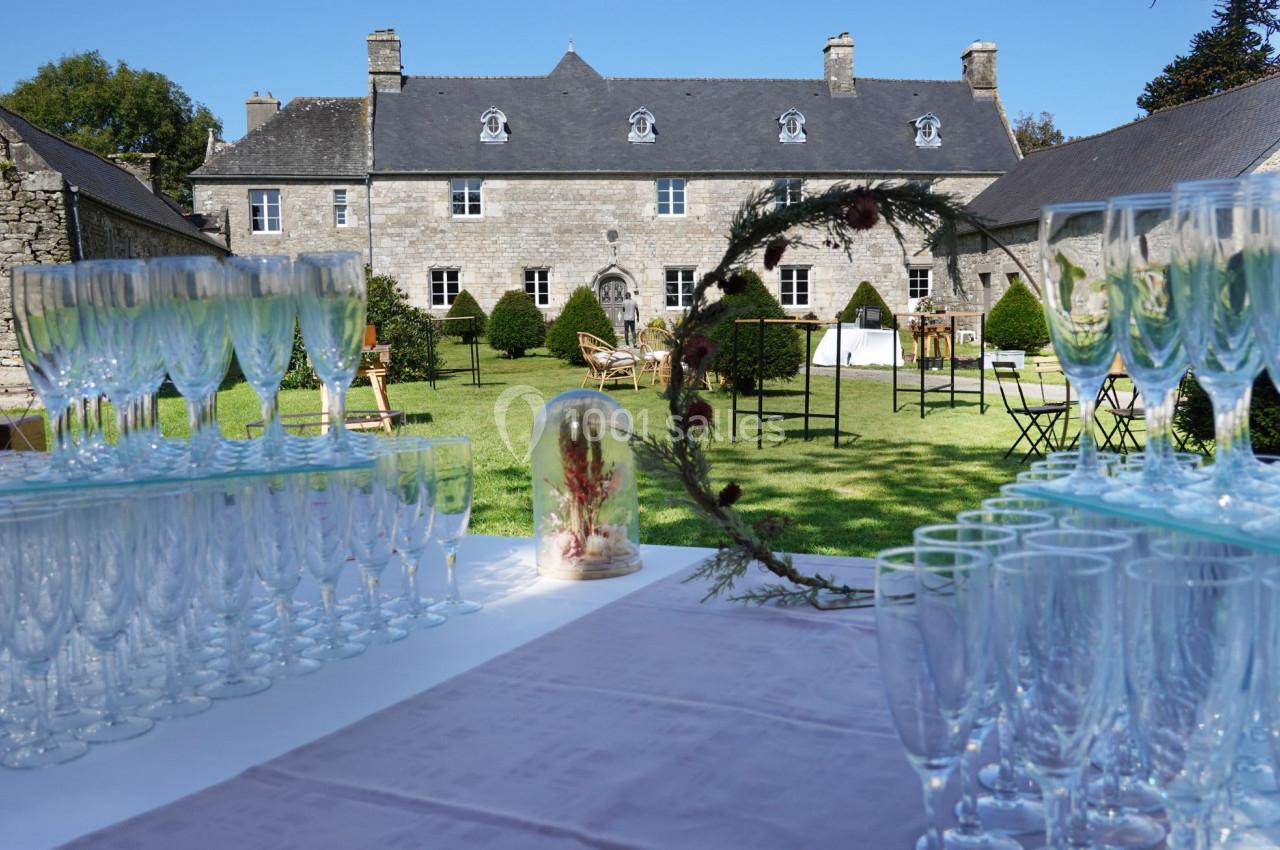 Table dressée avec des verres en cristal devant un manoir en pierre entouré d'un jardin verdoyant.