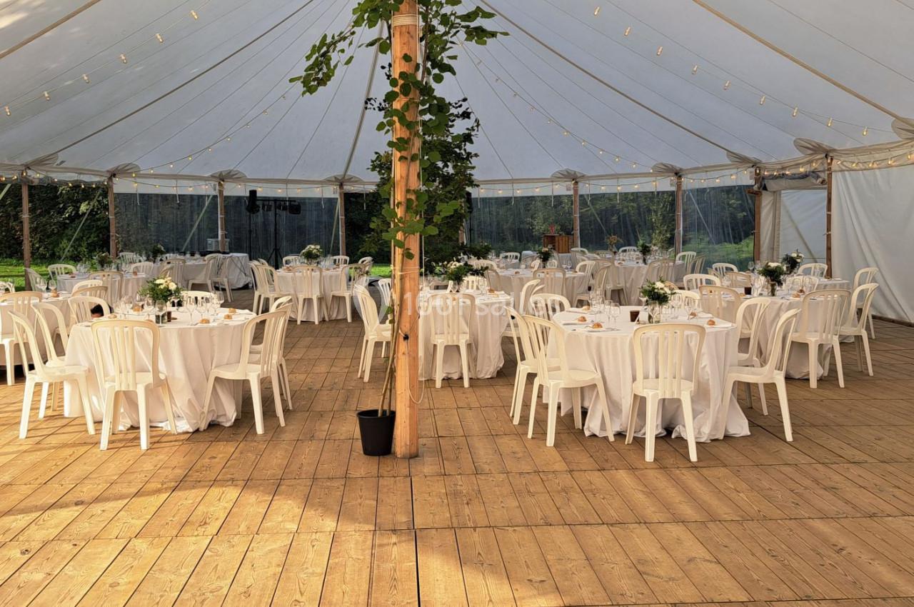 Intérieur d'une tente de réception avec des tables rondes dressées, des chaises blanches et une décoration végétale.