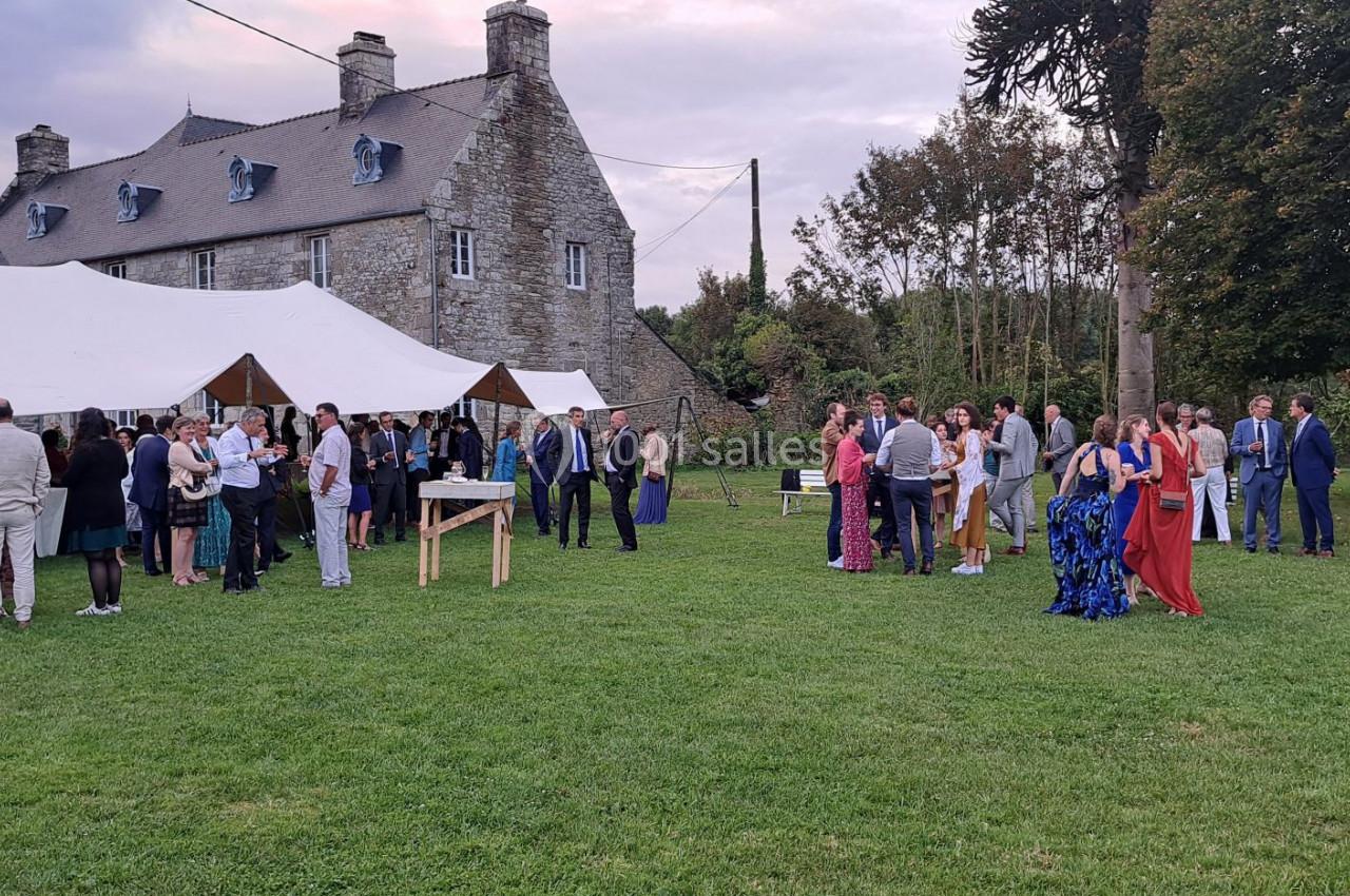 Groupe de personnes rassemblées dans un jardin devant une grande maison en pierre et des tentes blanches.