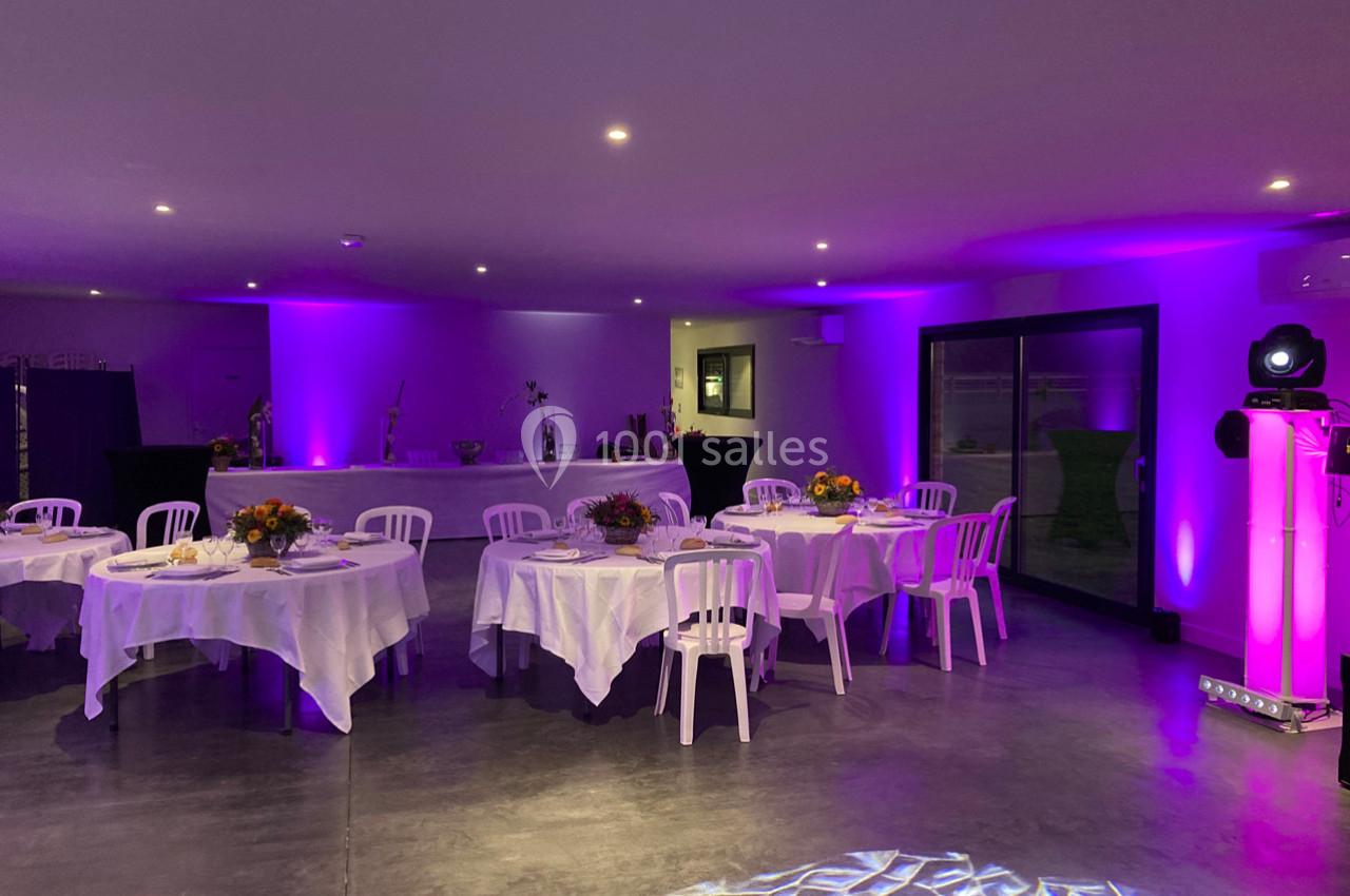 Salle de réception décorée avec des tables rondes, nappes blanches, chaises et éclairage violet tamisé.