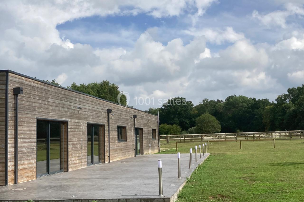 Bâtiment en bois avec une terrasse en béton, entouré de pelouses et de clôtures dans un environnement rural.