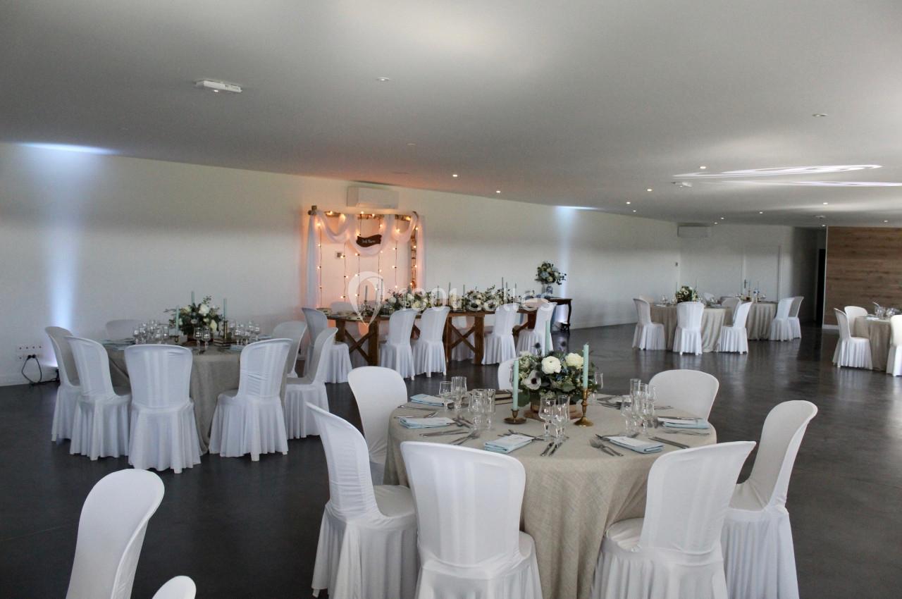 Salle de réception décorée avec des tables rondes et rectangulaires, nappes blanches et arrangements floraux.