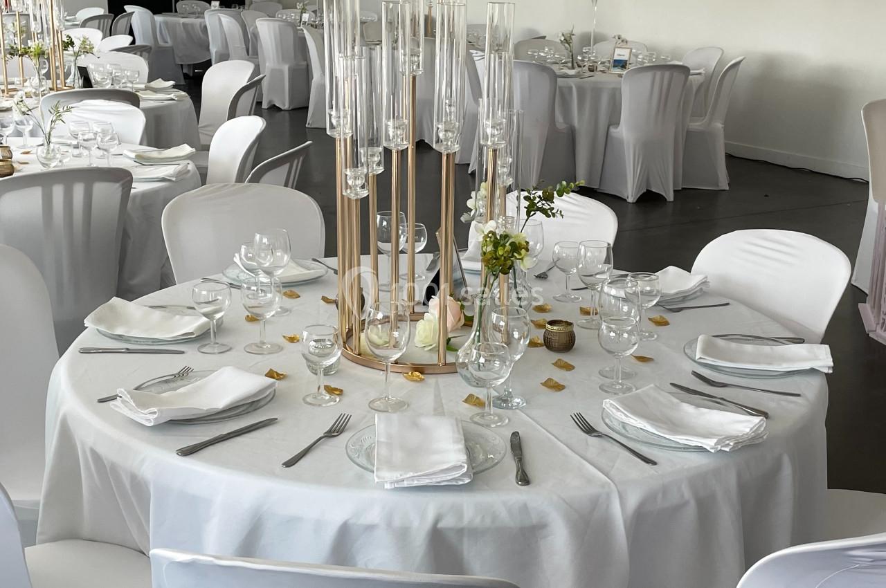 Table ronde élégamment dressée avec nappes blanches, serviettes pliées, verres, couverts et centre de table décoratif.