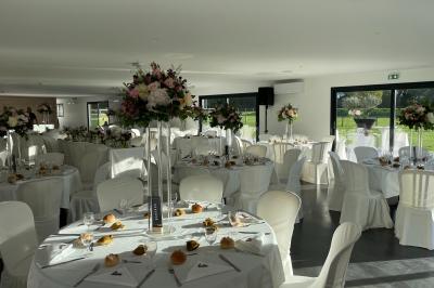Salle de réception lumineuse avec tables rondes décorées de nappes blanches, fleurs et vaisselle élégante.
