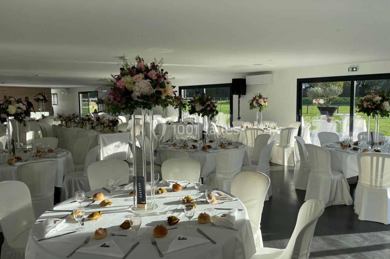 Salle de réception lumineuse avec tables décorées de nappes blanches, centres de table fleuris et vaisselle élégante.