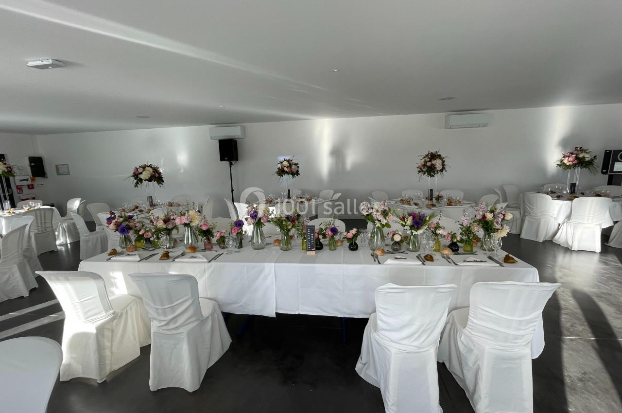 Salle de réception décorée pour un événement, avec des tables ornées de fleurs et des chaises recouvertes de housses…
