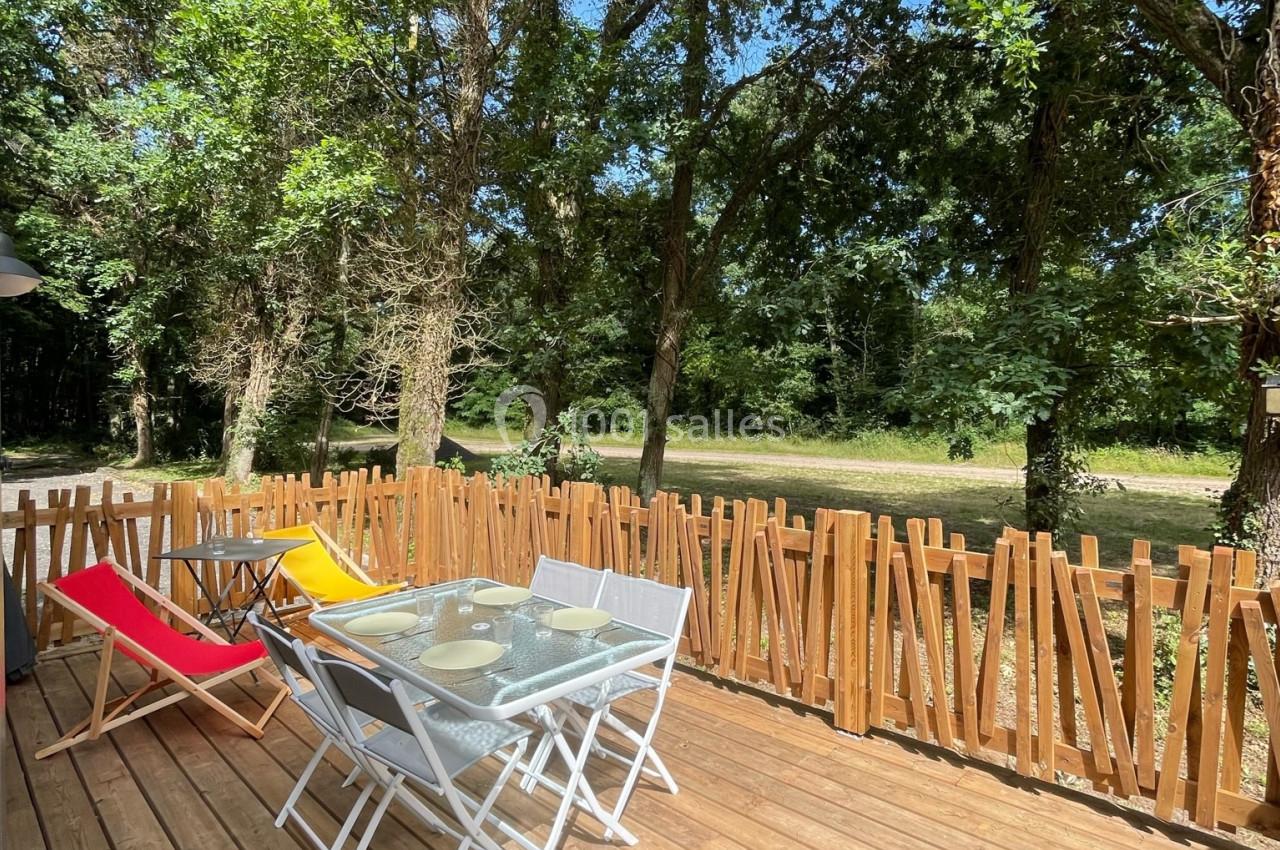 Terrasse en bois avec table, chaises blanches et transats colorés, entourée d'une clôture et bordée d'arbres.