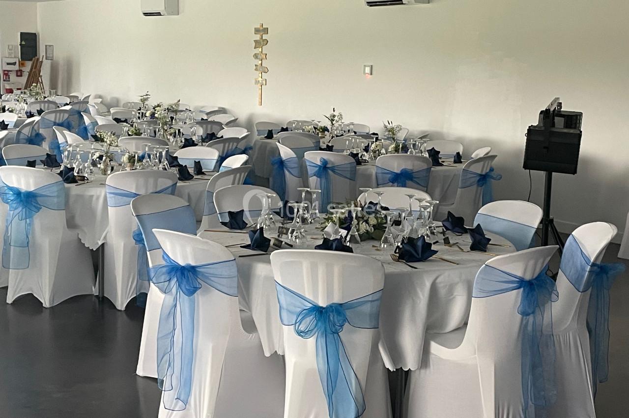 Salle de réception décorée avec des tables rondes, nappes blanches et chaises ornées de nœuds en tissu bleu.