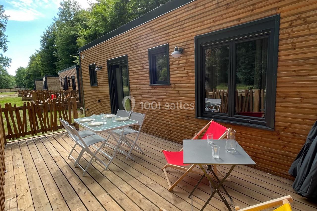 Terrasse en bois avec table, chaises et transats colorés, devant un logement en bois entouré de verdure.