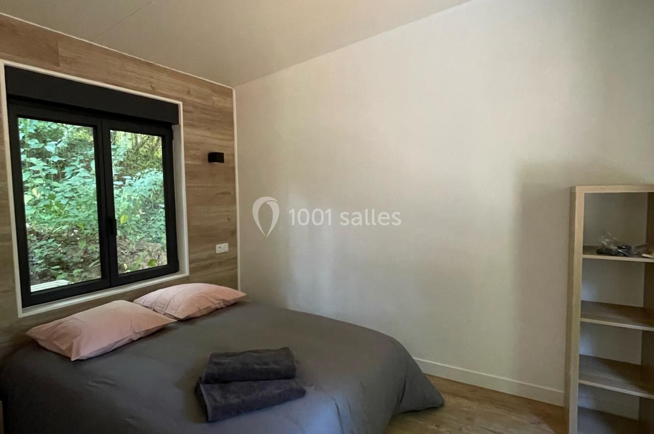 Chambre avec lit double, draps gris, oreillers roses, étagère en bois et fenêtre donnant sur un paysage boisé.