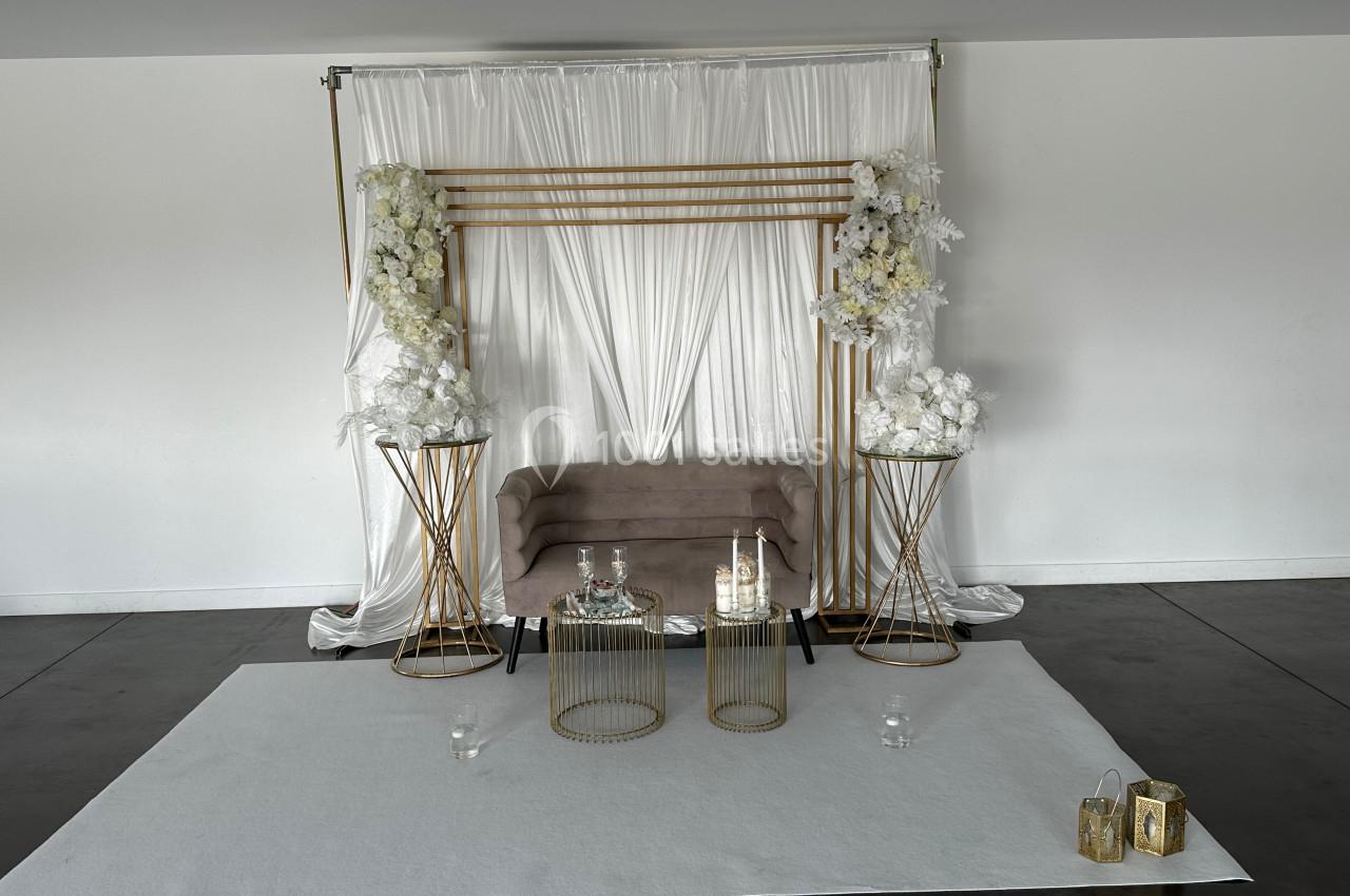 Décor minimaliste avec un canapé beige, des fleurs blanches sur des supports dorés et un fond en tissu blanc.