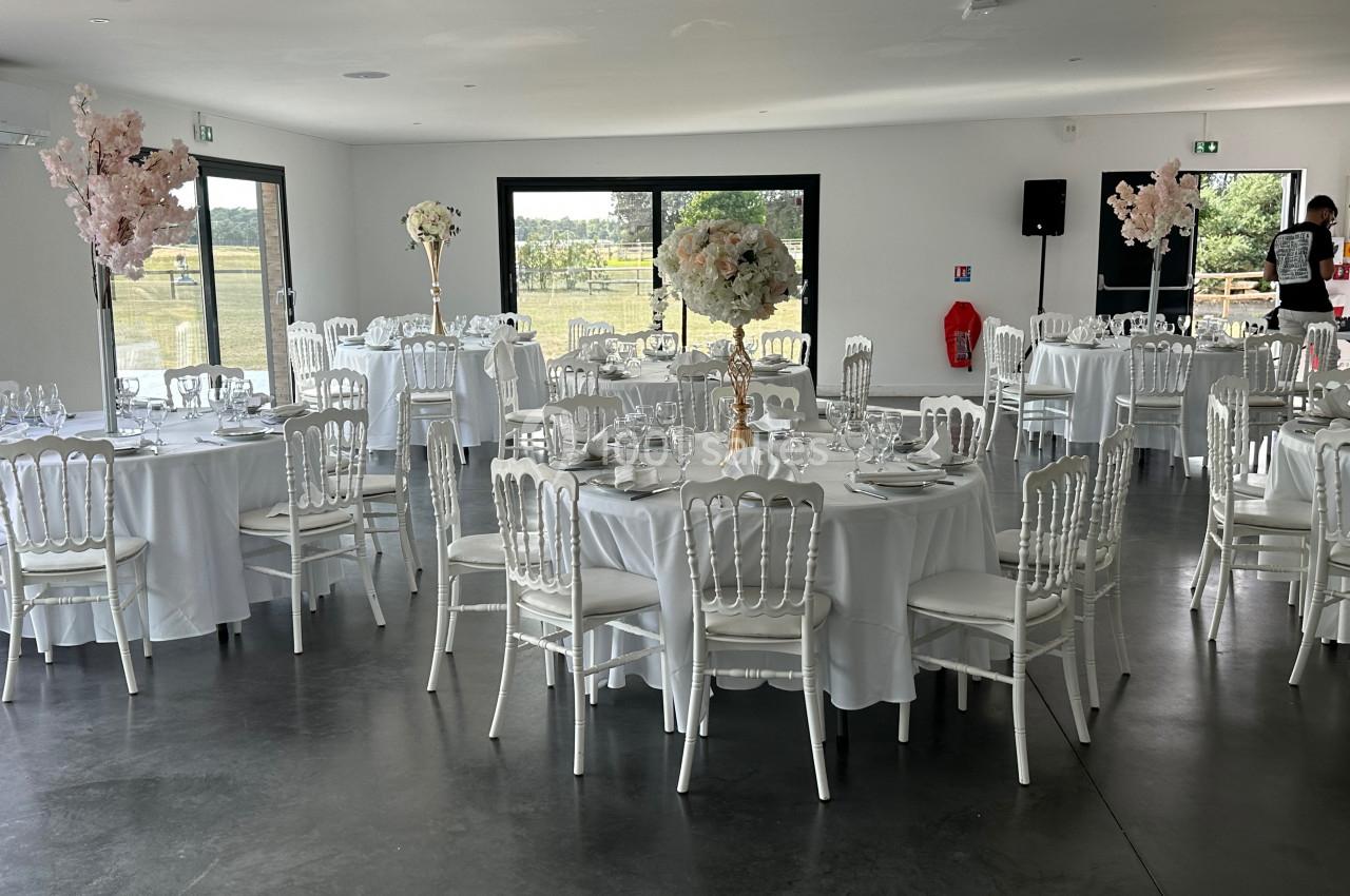 Salle de réception lumineuse avec tables rondes dressées, nappes blanches et décorations florales élégantes.