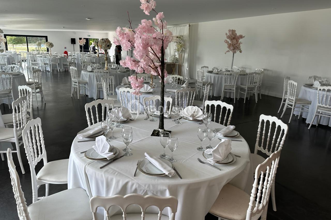 Salle de réception avec tables rondes dressées, nappes blanches, serviettes pliées et décorations florales roses.