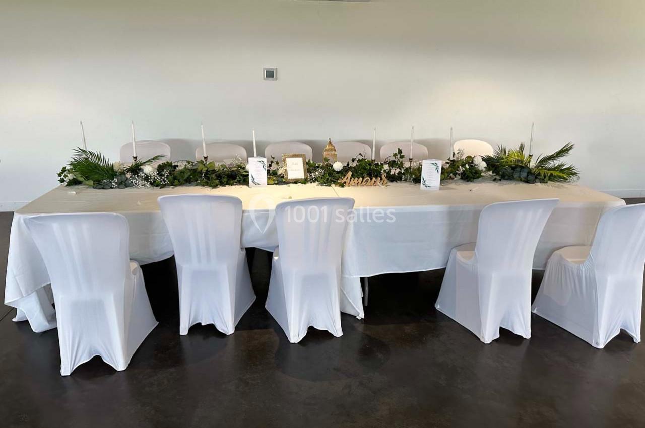 Table décorée avec une nappe blanche, des chaises houssées, un centre de table végétal et des bougies dans une salle…
