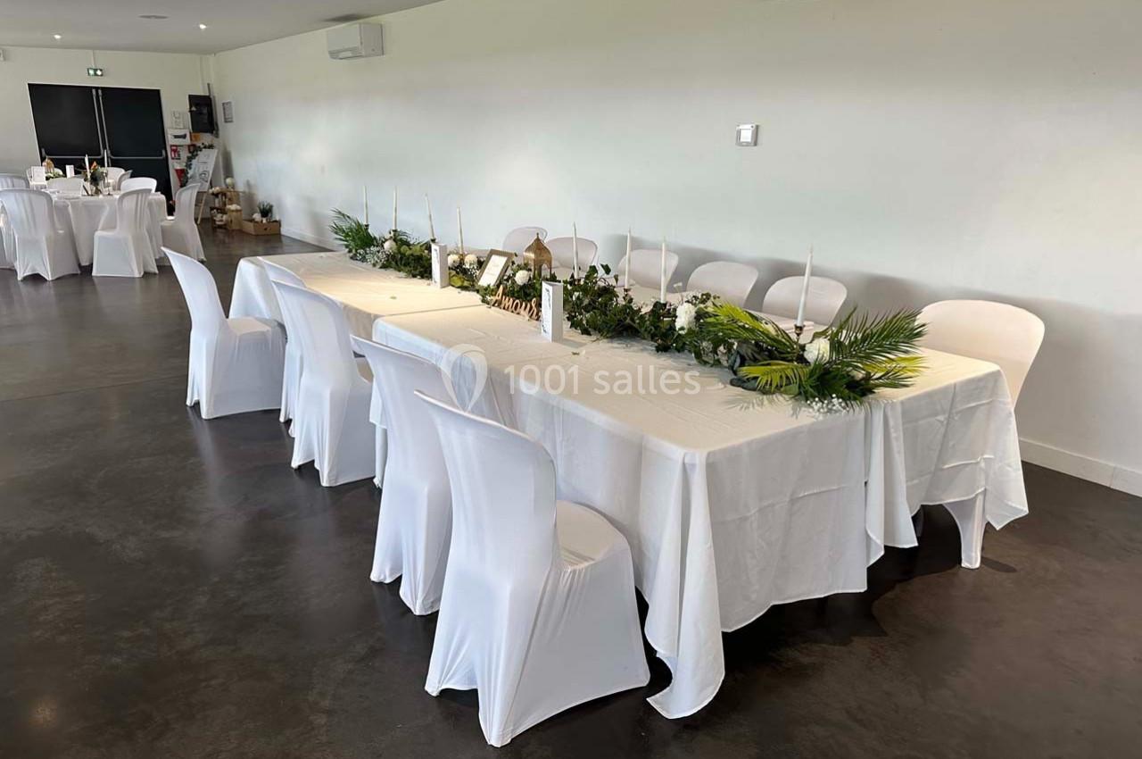 Grande table rectangulaire décorée de feuillages, bougies et nappes blanches, entourée de chaises également blanches.