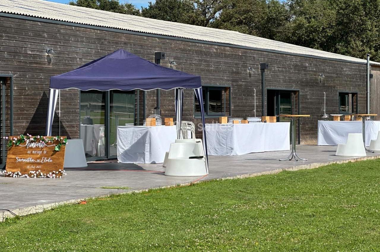 Terrasse avec tables dressées sous un barnum violet, devant un bâtiment en bois, entourée de pelouse.