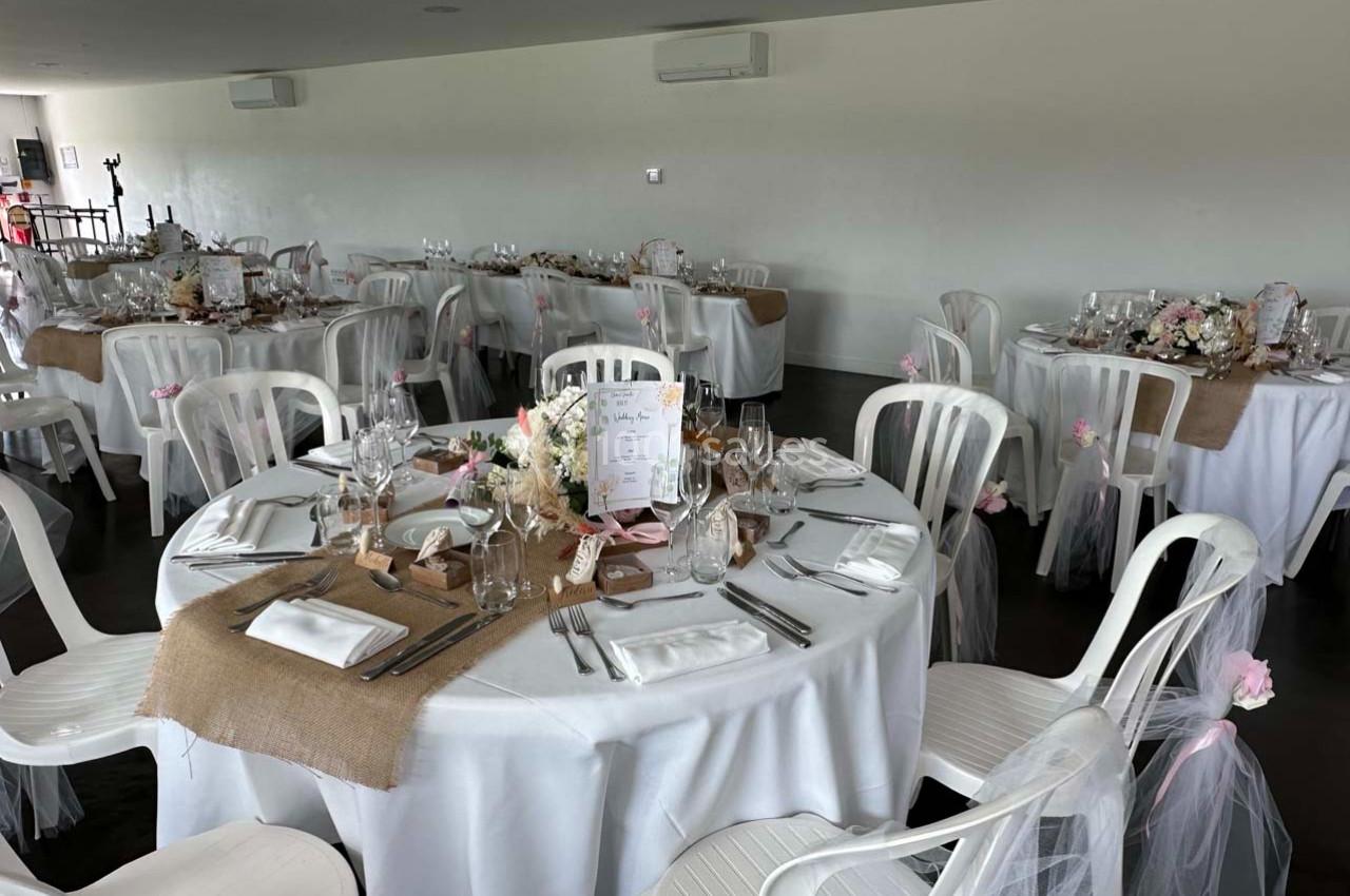 Salle de réception décorée avec des tables rondes dressées, nappes blanches, chemins de table en toile de jute et chaises…