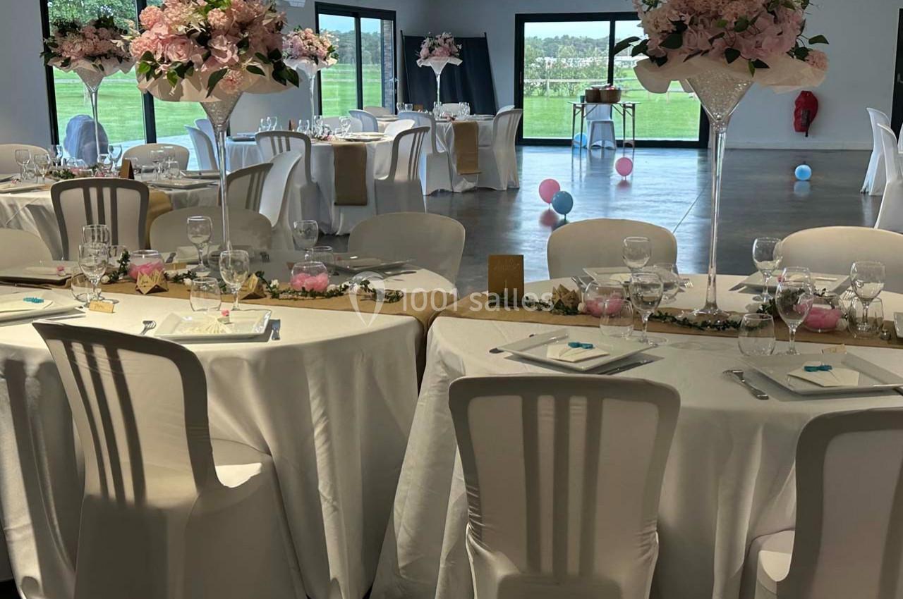 Salle de réception décorée pour un mariage avec tables rondes, nappes blanches, centres de table fleuris et baies vitrées.