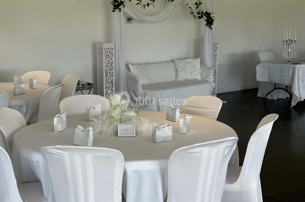 Salle décorée pour un événement avec des tables rondes couvertes de nappes blanches et une arche ornée de voilages.