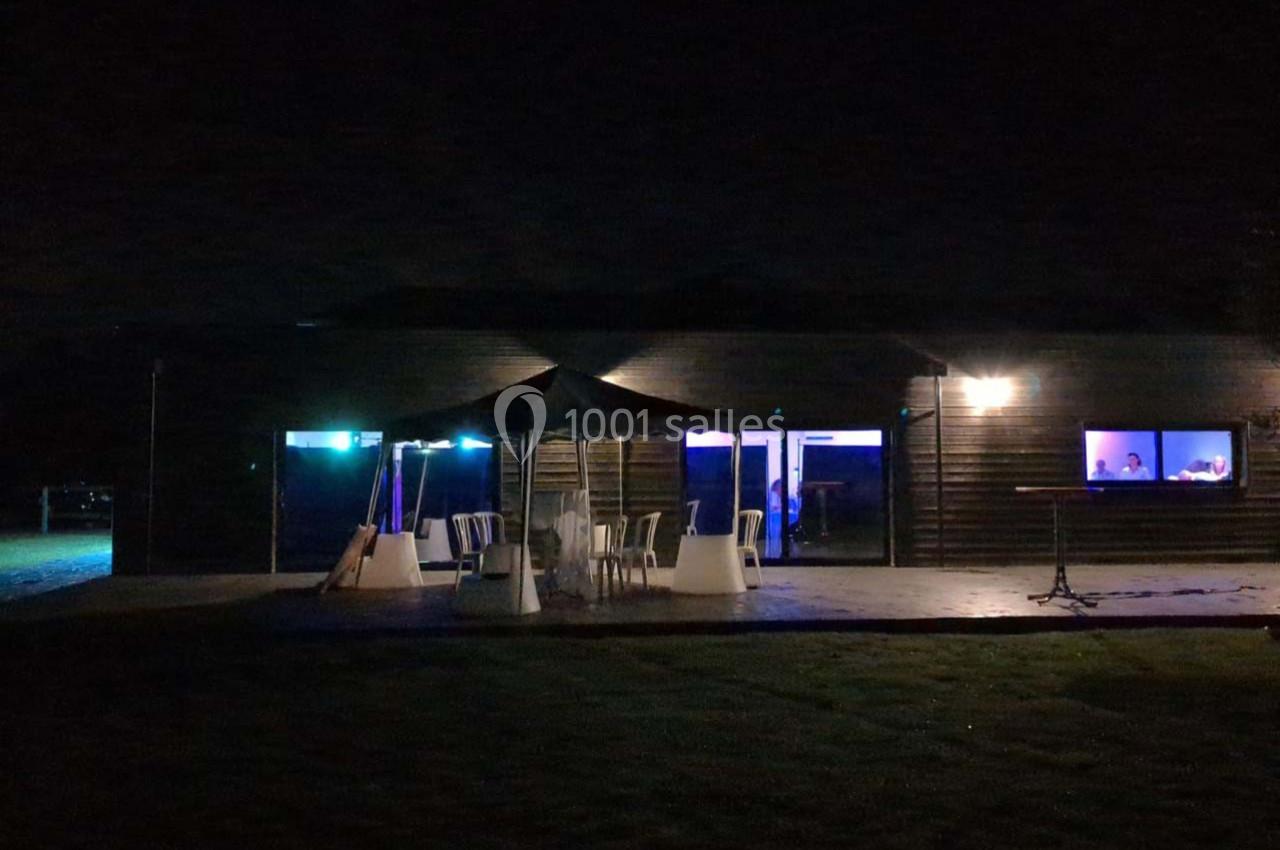 Terrasse extérieure sombre avec des chaises et une table sous un parasol, éclairée par des lumières bleues et jaunes.