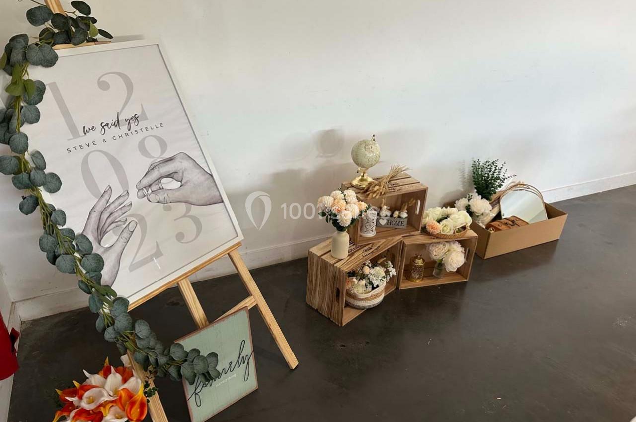 Décoration de mariage avec un chevalet illustré, des fleurs, des caisses en bois et des accessoires sur un sol sombre.