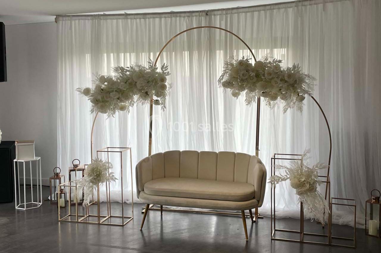 Canapé beige entouré d'arcs dorés décorés de fleurs blanches, devant un rideau blanc translucide.