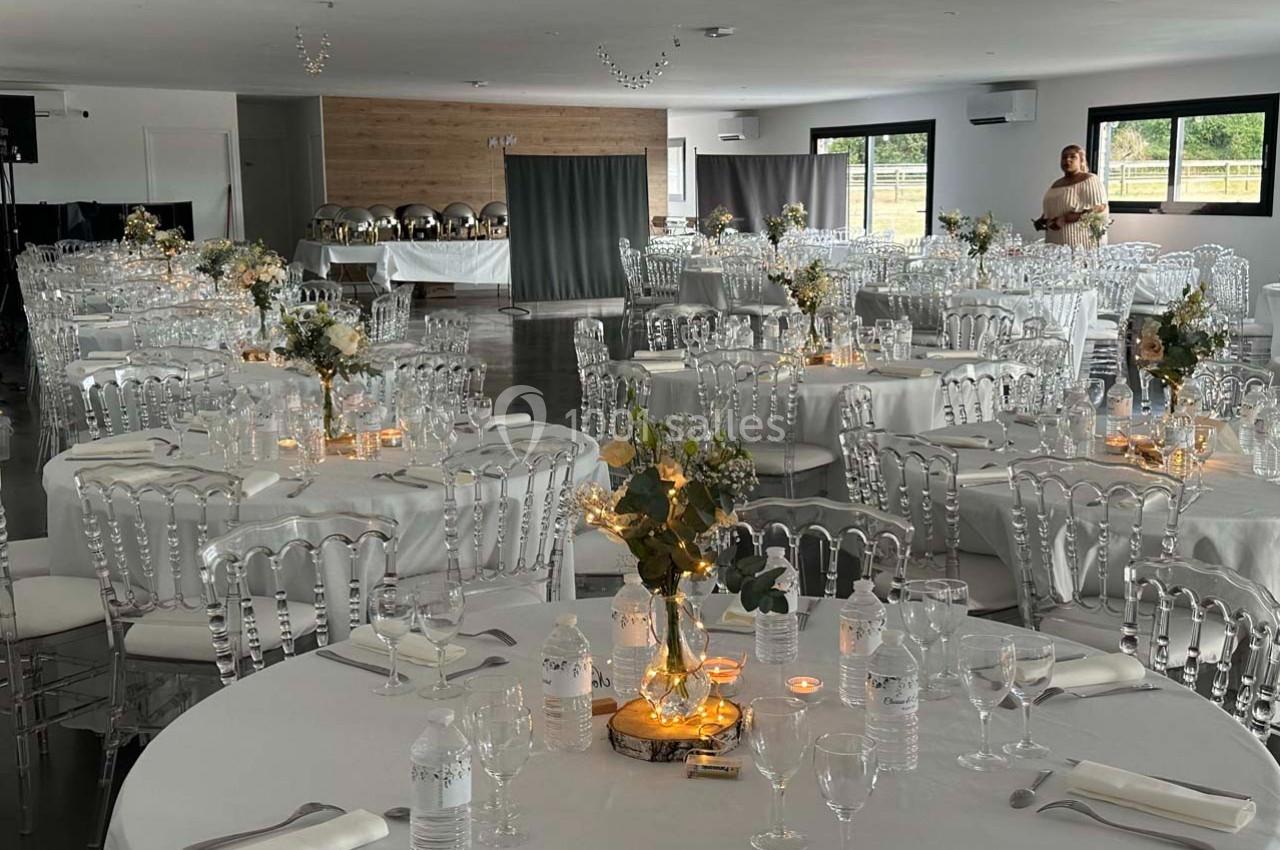 Salle de réception décorée avec des tables rondes, nappes blanches, chaises transparentes et centres de table fleuris.