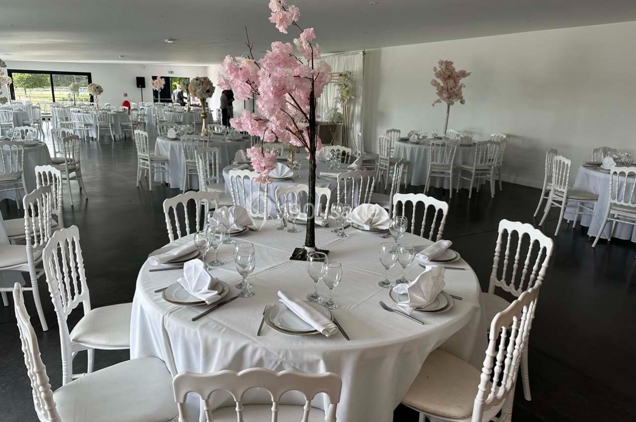 Salle de réception avec tables rondes dressées, nappes blanches et centres de table décorés de branches fleuries roses.