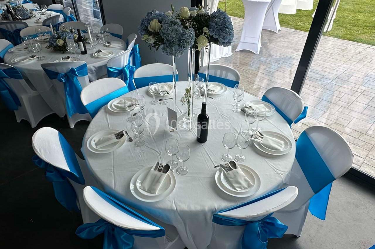 Tables rondes décorées de nappes blanches et de nœuds bleus, avec des centres de table floraux et des couverts disposés.