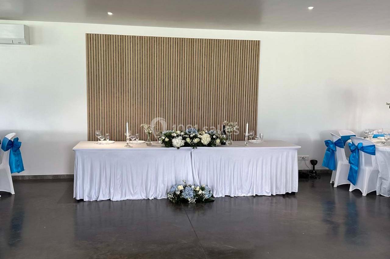 Table décorée pour un mariage avec nappes blanches, fleurs blanches et bleues, et chaises ornées de nœuds bleus.