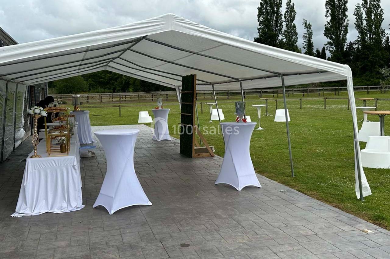 Tente blanche installée sur une terrasse pavée, avec des tables hautes habillées de nappes blanches et un espace vert en…