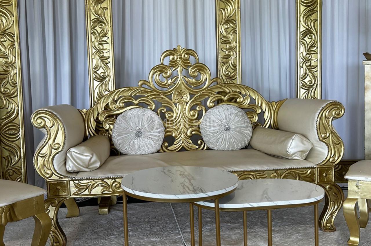 Canapé doré orné de motifs baroques, accompagné de coussins décoratifs et de tables basses en marbre blanc.
