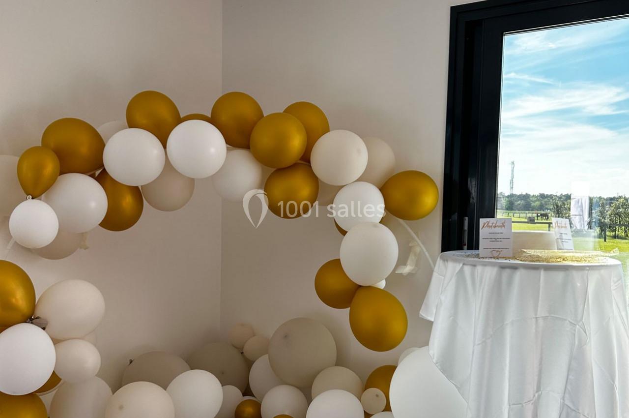 Arche de ballons blancs et dorés près d'une table drapée de blanc, avec vue sur un paysage extérieur ensoleillé.