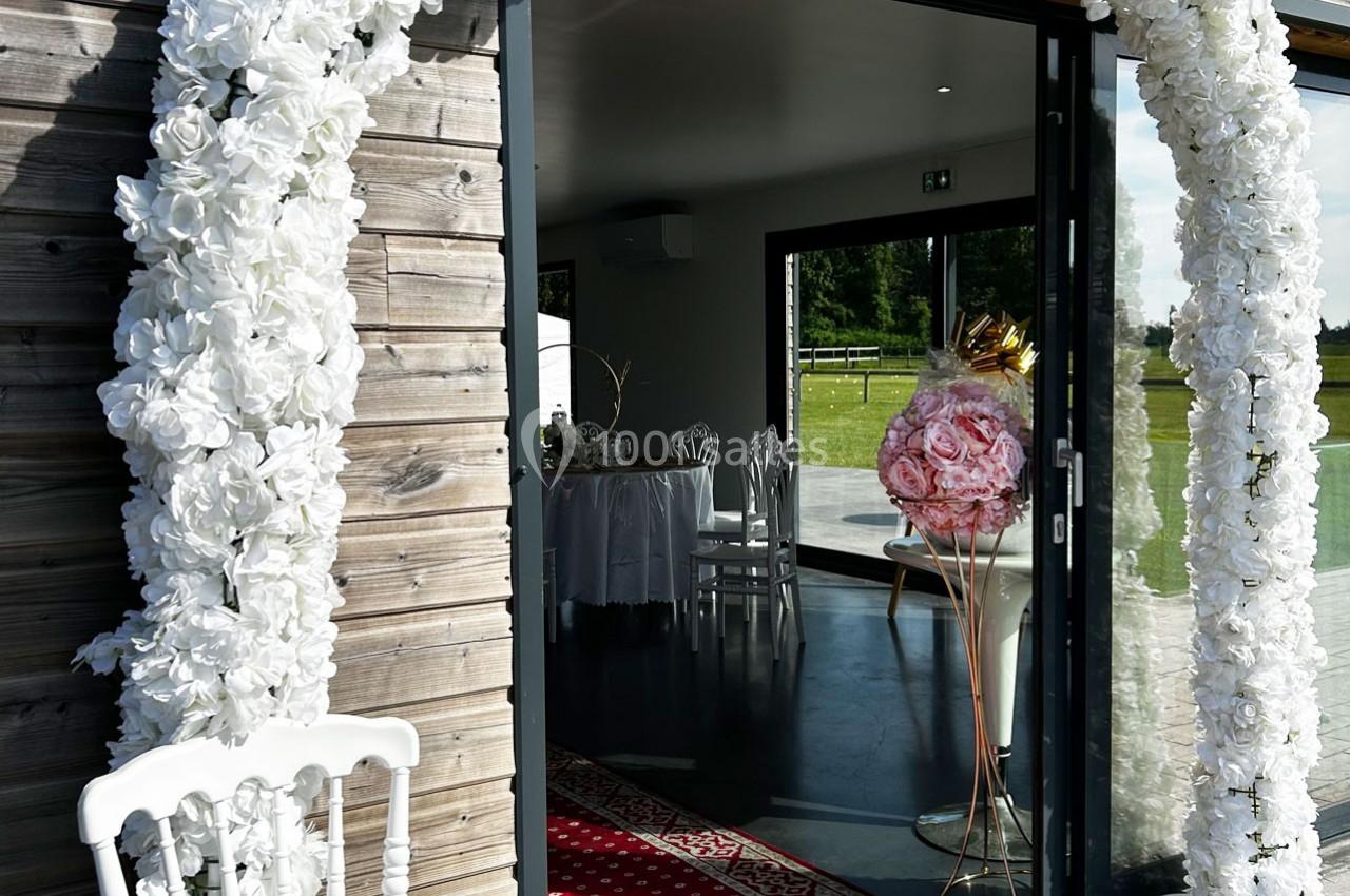 Entrée décorée de fleurs blanches menant à une salle de réception lumineuse avec des chaises et une table dressée.